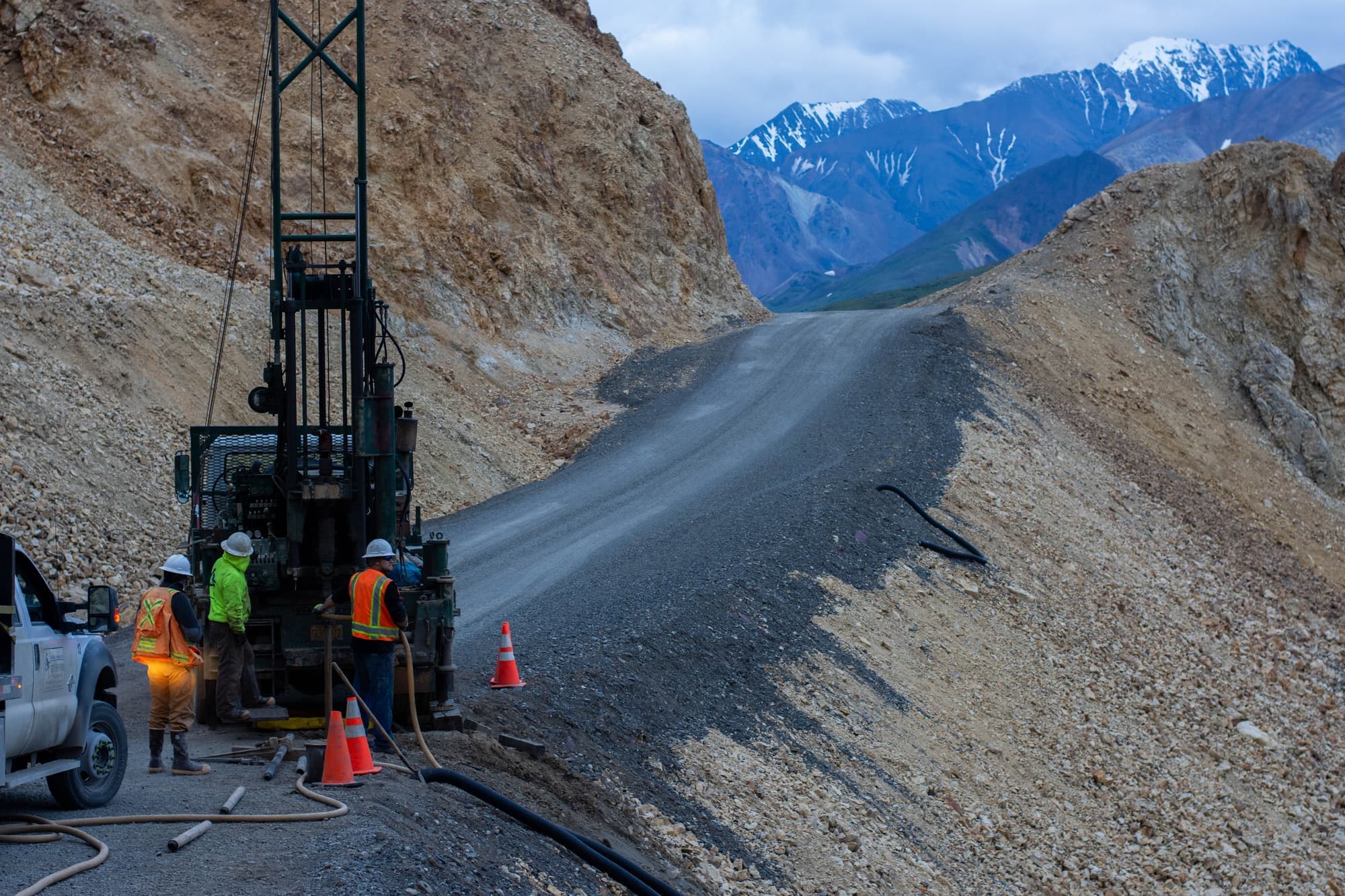 Core drilling crew working a rig on a mountain road, snow-capped peaks behind